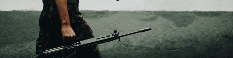 A soldier in camouflage walking with a rifle against a textured wall, day.